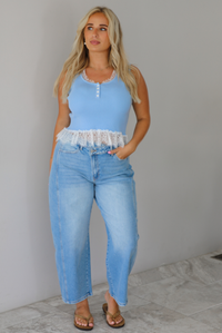 A woman wearing a light blue sleeveless top with a white lace hem and light blue high-waisted cropped jeans, standing indoors against a white wall with white marble flooring. 
