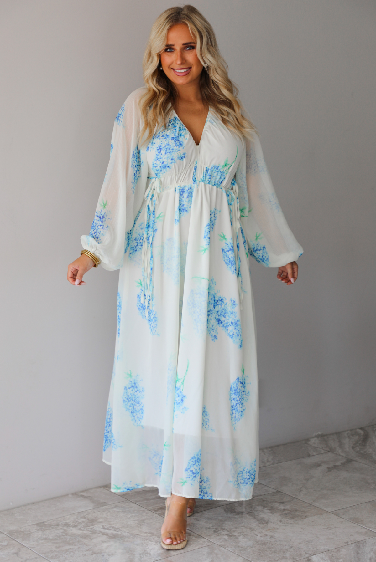 A woman modeling a white maxi dress with a blue floral print and a sheer, flowing overlay, standing indoors against a white wall with white marble flooring. 