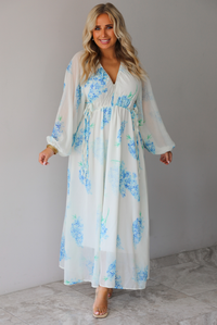A woman modeling a white maxi dress with a blue floral print and a sheer, flowing overlay, standing indoors against a white wall with white marble flooring. 