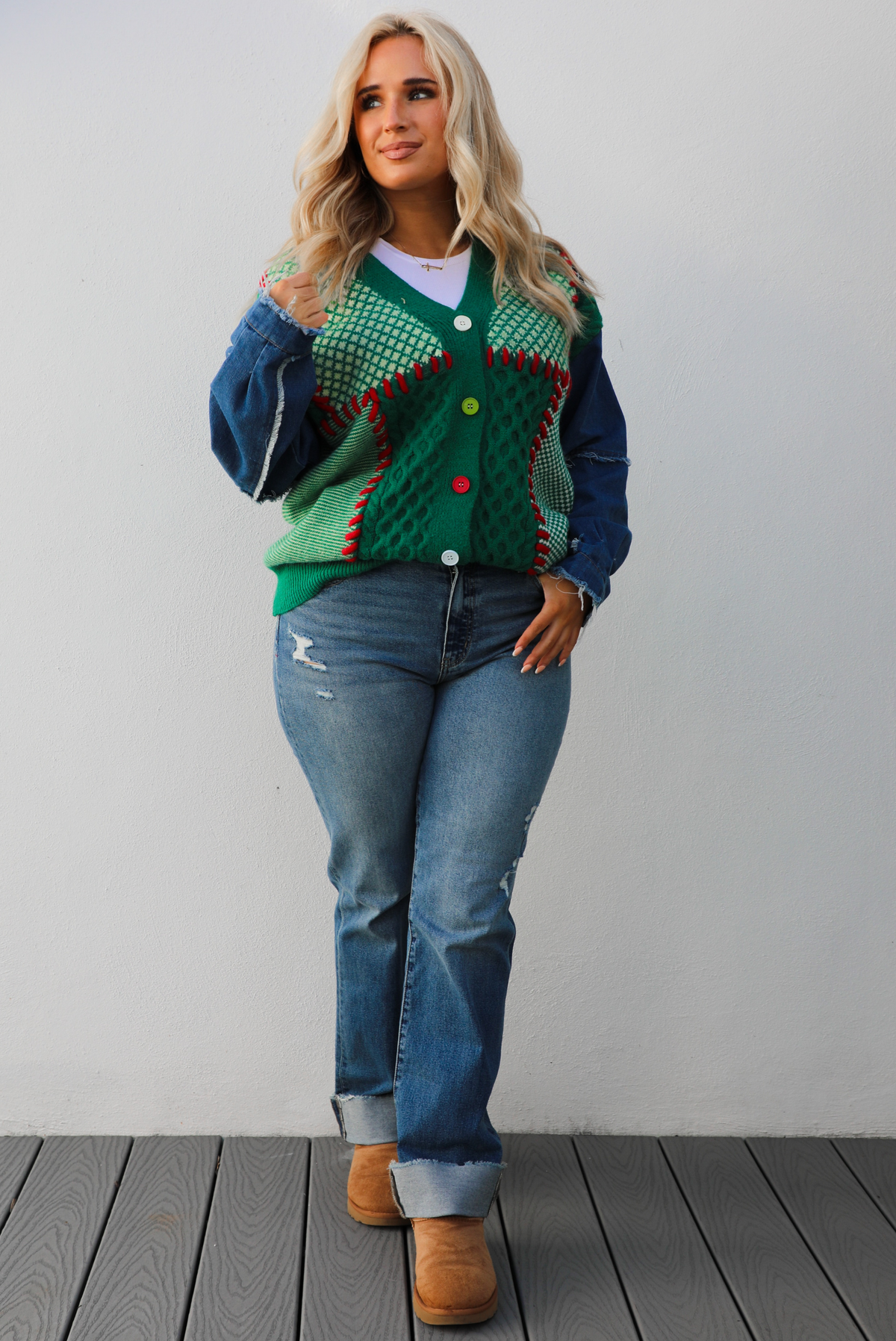 Woman model wearing a green and multi-colored cardigan with denim sleeves, medium wash denim jeans, and tan uggs. The photo is taken against a white wall outdoors.
