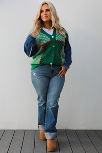 Woman model wearing a green and multi-colored cardigan with denim sleeves, medium wash denim jeans, and tan uggs. The photo is taken against a white wall outdoors.