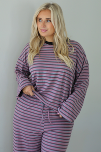 A woman wearing a purple matching set with a black and blue striped pattern featuring an oversized long sleeved top and high waisted flared hem pants, standing indoors against a white wall. The photo shows a closeup of the top, which has a thick black neckline detail. 