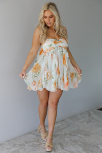 Woman wearing a white, strapless, babydoll cut dress that has an orange and green floral print pattern and flowy skirt indoors against a white wall with white marble flooring. 