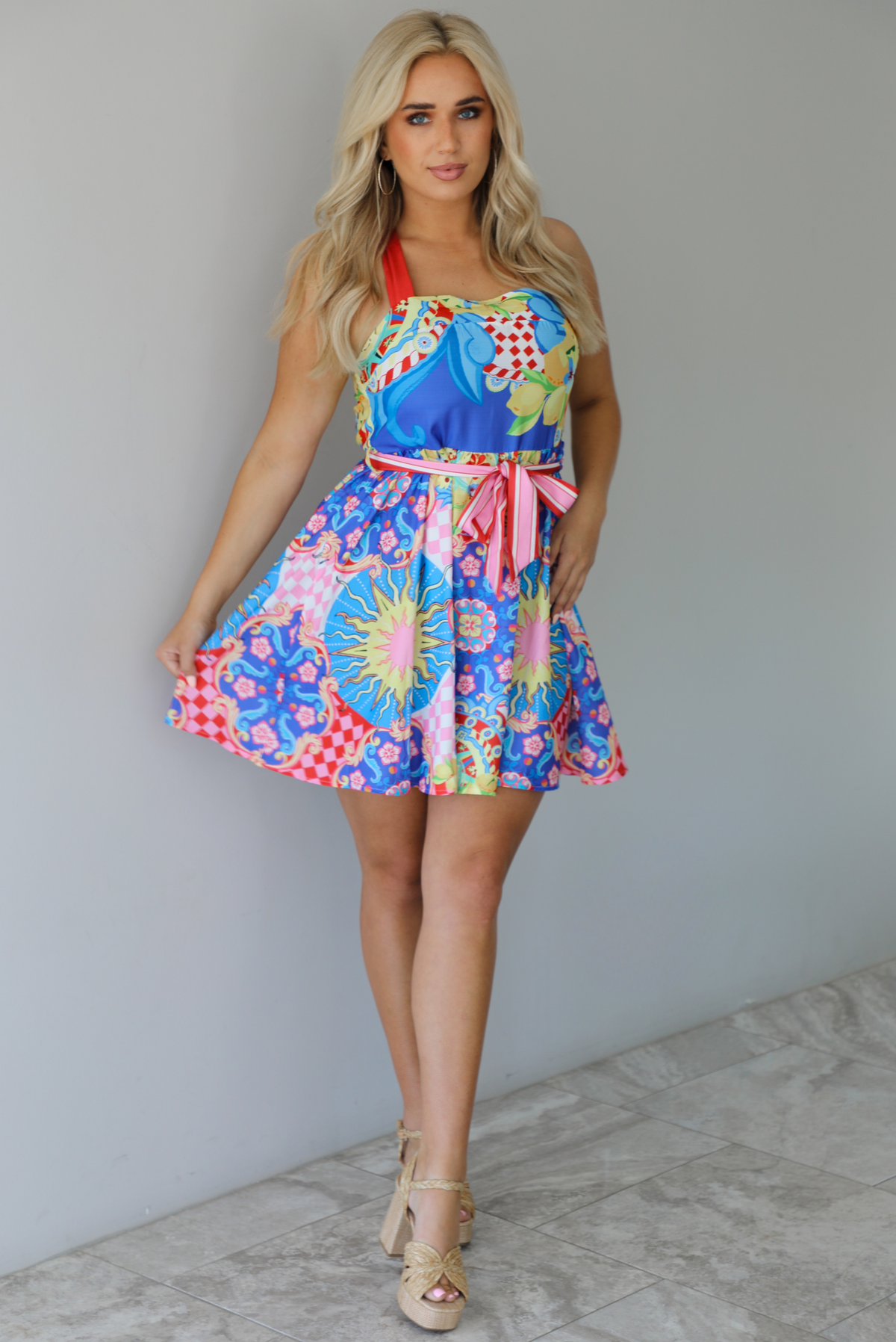 Woman wearing a multi-colored and multi-patterned mini dress with thick red straps, a cinched waistline with a removable tie, and a flowy skirt indoors against a white wall with white marble flooring. 
