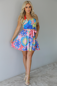 Woman wearing a multi-colored and multi-patterned mini dress with thick red straps, a cinched waistline with a removable tie, and a flowy skirt indoors against a white wall with white marble flooring. 