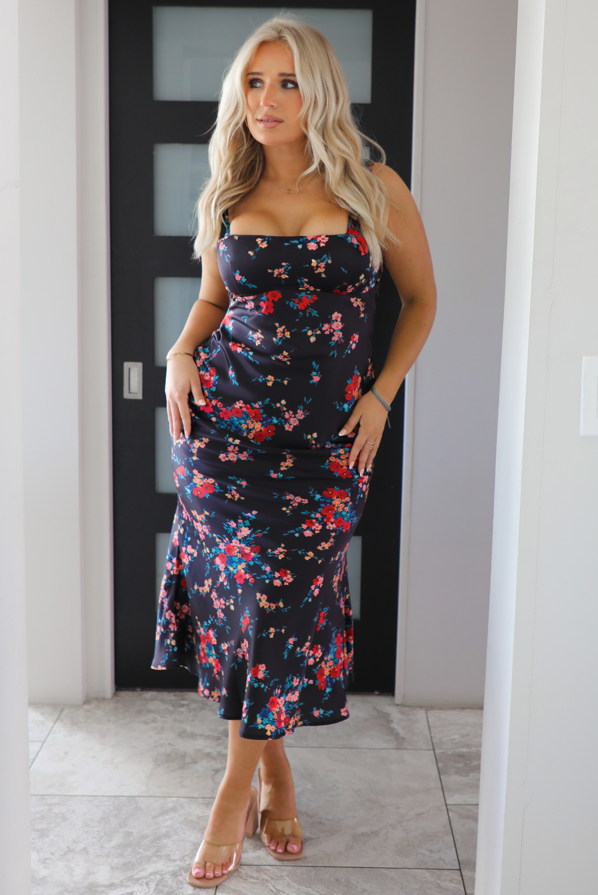 Woman model wearing a navy dress with a multi-colored floral print. The dress is a satin material, is a midi length, and is curve-hugging. The photo is taken inside a modern white and black interior. 