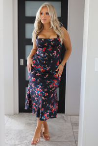 Woman model wearing a navy dress with a multi-colored floral print. The dress is a satin material, is a midi length, and is curve-hugging. The photo is taken inside a modern white and black interior. 