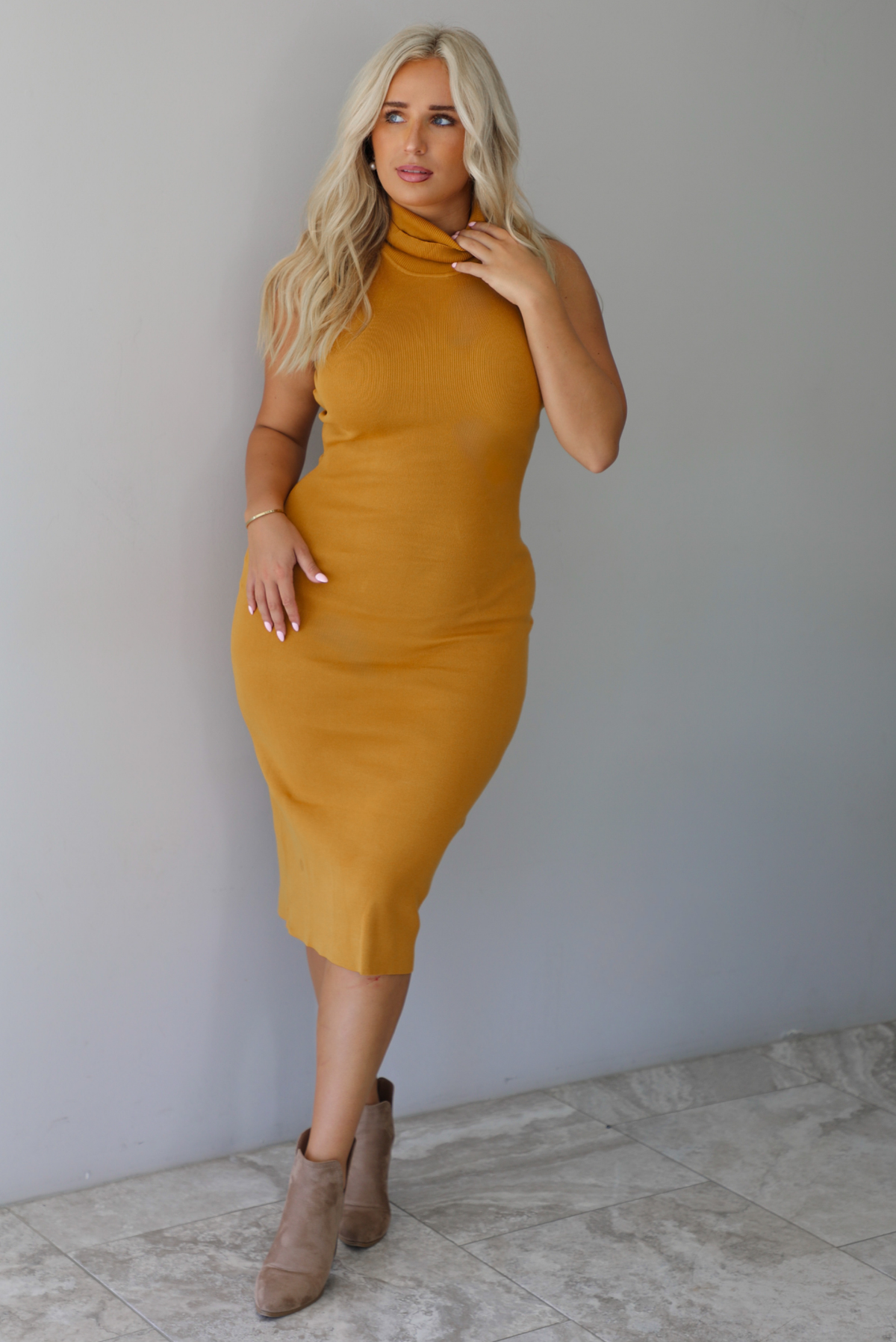 A woman wearing a marigold sleeveless midi dress with a high, turtle neckline, ribbed textured material, and is fitted, standing indoors against a white wall with white marble flooring. 