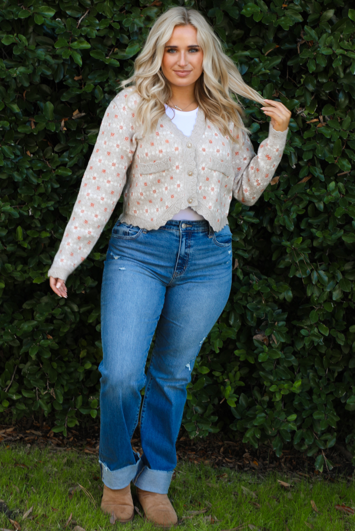 Woman model wearing a grey cardigan that has an orange and white floral print pattern throughout, long sleeves, an asymmetrical hem, and functional button closures in the front, with a long sleeved white top, medium wash denim jeans, and brown shoes in front of a green hedge outdoors. 