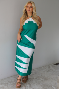 Woman wearing a green and white maxi dress with a halter neckline indoors against a white wall with white marble flooring. 