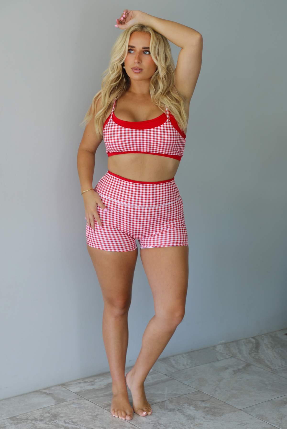 A woman modeling a red and white gingham two-piece athletic set with solid red trim indoors against a white wall with white marble flooring. 