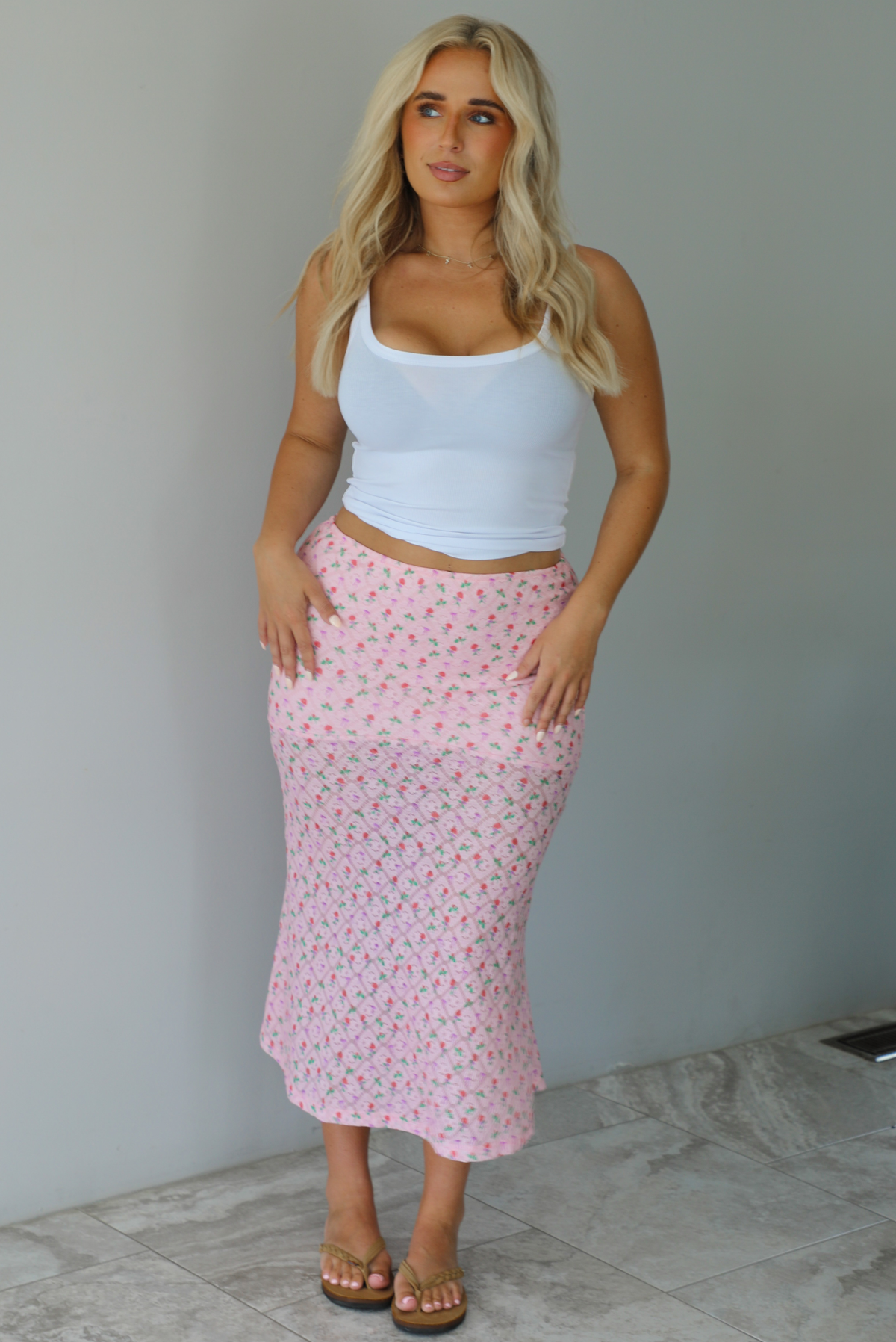 Woman wearing a white tank and pink midi length skirt that has a repeating floral pattern throughout indoors against a white wall with white marble flooring. 