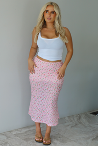Woman wearing a white tank and pink midi length skirt that has a repeating floral pattern throughout indoors against a white wall with white marble flooring. 