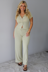 A woman wearing a two piece set consisting of a collared, button-down, short sleeve top and high waisted, drawstring, wide leg pants, standing indoors against a white wall with white marble flooring. 