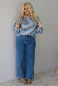 A woman modeling a light blue and navy striped long-sleeve sweater with a white ruffled collar, paired with wide-leg blue jeans and brown boots, standing indoors against a white wall with white marble flooring. 