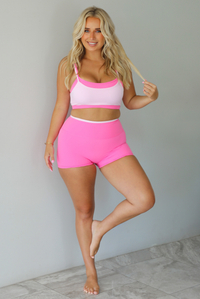 A woman modeling a bright pink and light pink two-piece activewear set consisting of a sports bra and matching high-waisted shorts, standing indoors against a white wall with white marble flooring. 