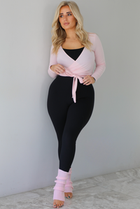 Woman wearing a pink cropped wrap style fitted top over a black fitted jumpsuit. The outfit is paired with pink leg warmers. The photo is taken indoors against a white wall with white marble flooring. 