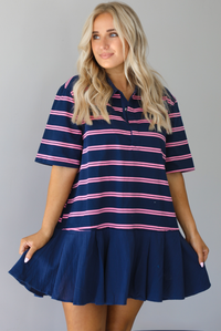 Woman wearing a navy dress with a pink striped design throughout, collared neckline, short sleeves, and a flowy bottom hem that's a different texture than the rest of the dress and is solid navy. The photo is taken indoors against a white wall.
