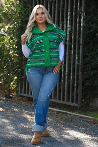 Woman model wearing a green, black, and white striped vest that has a sherpa texture to it, functional button closures in the front, and tie closures on either side, It is paired with a white long sleeved top, a medium wash pair of denim jeans, and tan ugg boots. The photo is taken outdoors with a metal gate and greenery in the back.