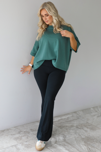 Woman in a teal oversized t-shirt paired with black leggings that flare slightly at the hem. The photo is taken indoors against a white wall with white marble flooring. 