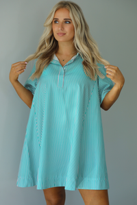 Woman wearing a green and pink striped dress that has a collared neckline, functional quarter button closures in the front, and a flowy skirt. The photo is taken indoors against a white wall.