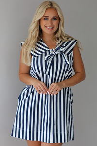 Woman wearing a navy and white striped dress with a bow detail at the neckline indoors against a white wall. The photo shows a closeup of the dress.