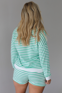 Woman wearing a green and white striped set featuring a v-neck long sleeved top and shorts indoors against a white wall. The photo shows the back of the set. 