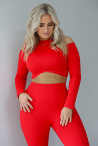 Woman wearing a two piece athletic set that has a cropped top with long sleeves, cold shoulder cut out details, and a cropped cut, with high waisted, fitted leggings. The photo shows a closeup of the cropped top. The photo is taken indoors against a white wall with white marbled flooring.