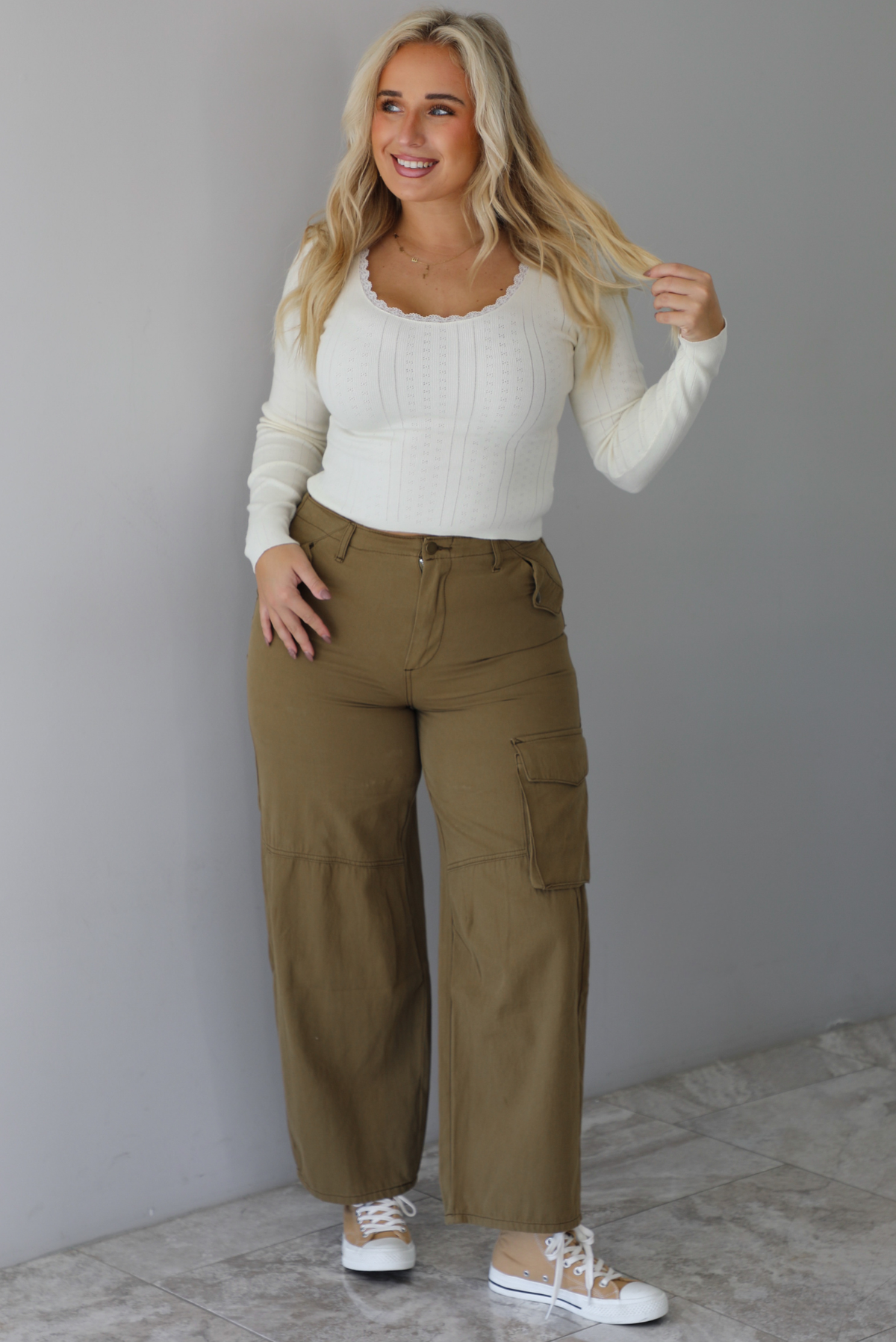 A woman wearing a cream knit long sleeved top and high waisted olive-khaki cargo pants, standing indoors against a white wall with white marble flooring. 
