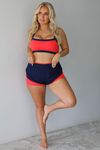 A woman modeling a two-piece athletic set with a bright coral-red and navy blue color-blocked design, standing indoors against a white wall with white marble flooring. 