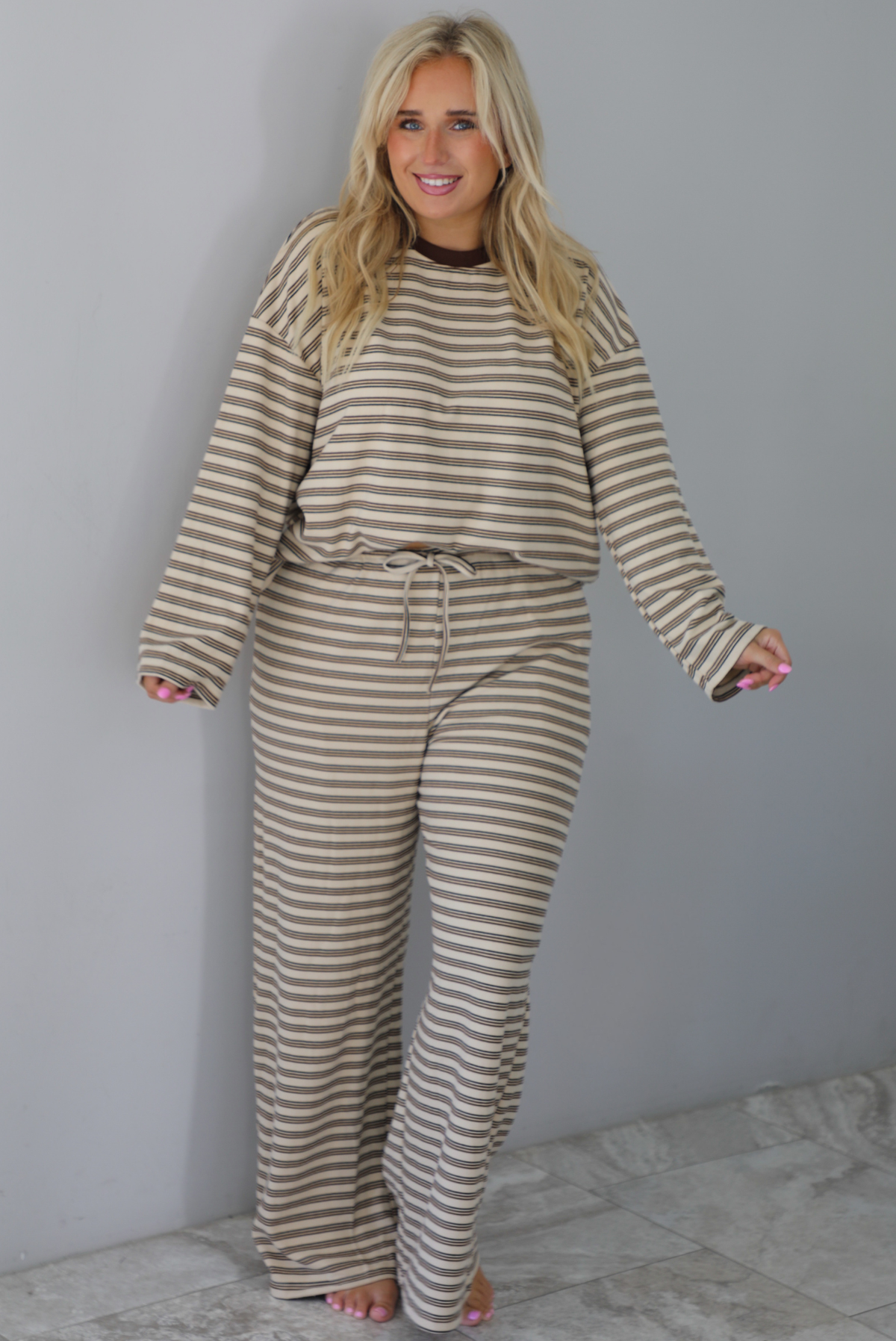 A woman wearing a sand colored matching two piece set with a brown striped pattern throughout featuring an oversized long sleeved top and oversized slouchy wide leg pants, standing indoors against a white wall with white marble flooring. 