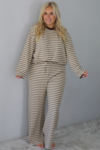 A woman wearing a sand colored matching two piece set with a brown striped pattern throughout featuring an oversized long sleeved top and oversized slouchy wide leg pants, standing indoors against a white wall with white marble flooring. 