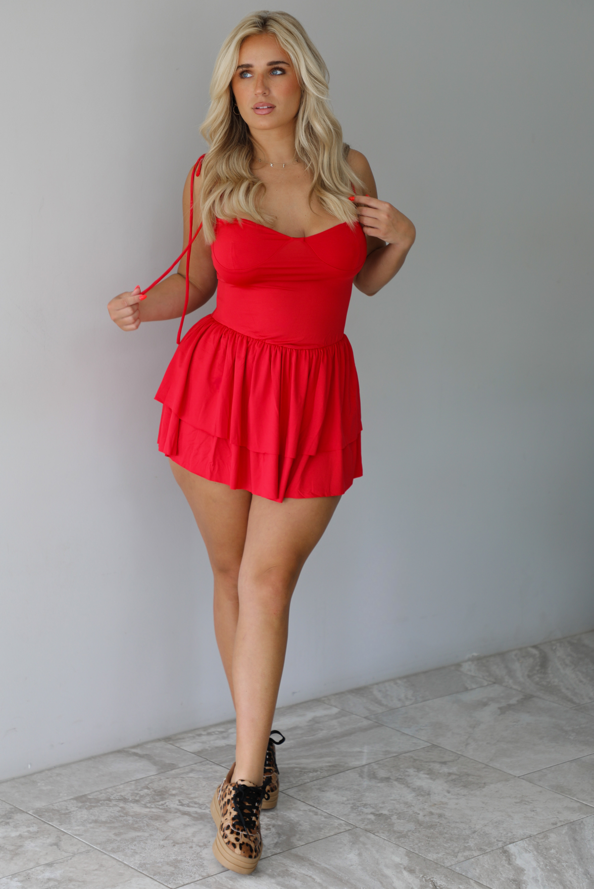 Woman wearing a red mini dress that has thin, spaghetti straps, a fitted bodice, and a flowy, ruffled skirt indoors against a white wall with white marble flooring. 