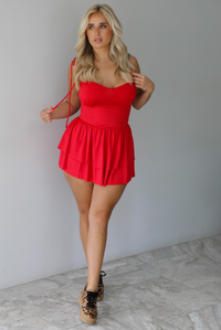 Woman wearing a red mini dress that has thin, spaghetti straps, a fitted bodice, and a flowy, ruffled skirt indoors against a white wall with white marble flooring. 