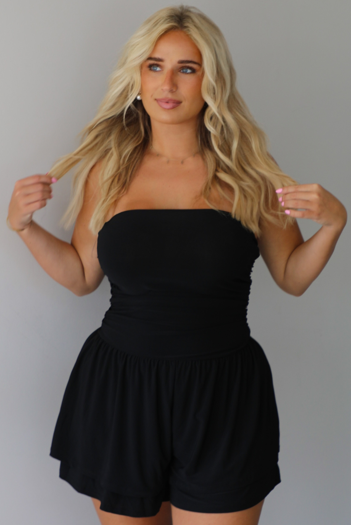 Woman wearing a black strapless romper with black boots indoors against a white wall.