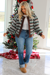 Woman model wearing a brown, green, and white Christmas themed sweater with multiple button closures in the front, slightly puffed sleeves, and a relaxed fit. The cardigan is paired with a white top underneath and medium wash denim jeans. The photo is taken inside a modern home with a decorated Christmas tree in the back. 