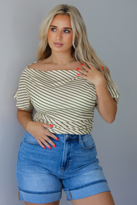 Woman wearing a pale yellow top with a taupe striped pattern with medium wash denim shorts indoors against a white wall.