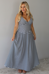 A woman modeling a sleeveless, V-neck maxi dress with a black and white horizontal stripe pattern, standing indoors against a white wall with white marble flooring. 