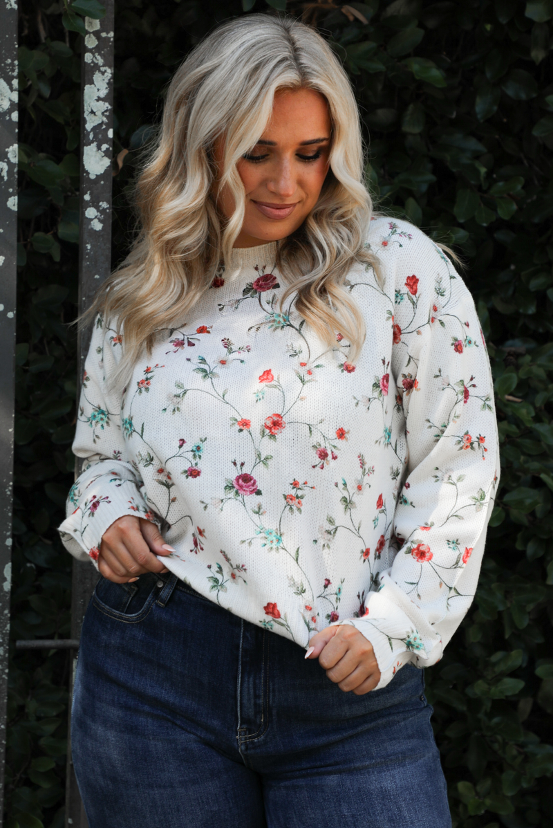 Woman model wearing an off-white sweater that has a multi-colored floral print pattern throughout with dark denim jeans. The photo showcases the length of the sweater and is close up. The photo is taken next to an iron gate outdoors with greenery in the back. 