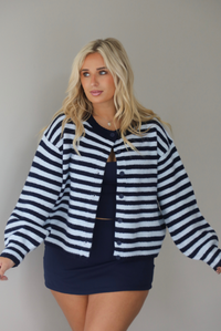 A woman wearing a navy and blue striped cardigan paired with a navy two piece set underneath standing indoors against a white wall.