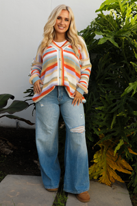 Woman model wearing an orange, lavender, yellow, muted green, blue, and white striped oversized cardigan with functional button closures in the front and an oversized fit with a light wash flared hem denim. The photo is taken outdoors against a white wall and greenery in the back. 