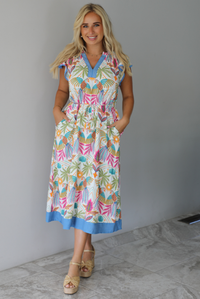 A woman wearing a short sleeved midi dress with a colorful tropical leaf print and light blue trim indoors against a white wall with white marble flooring. 