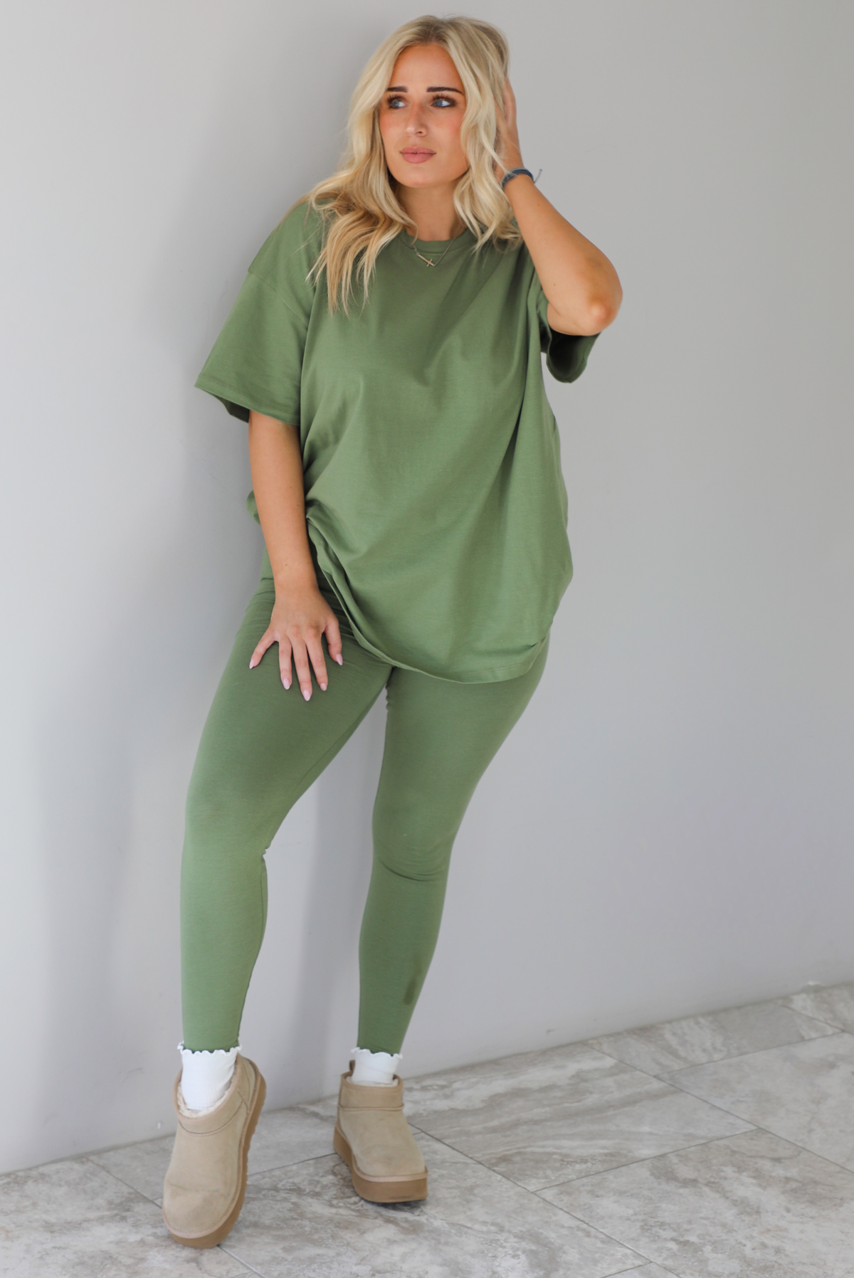 Woman wearing a green two piece set featuring an oversized t-shirt and fitted leggings. The set is paired with high white ankle socks and sand colored warm boots. The photo is taken indoors against a white wall with white marble flooring. 