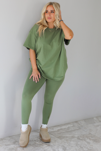 Woman wearing a green two piece set featuring an oversized t-shirt and fitted leggings. The set is paired with high white ankle socks and sand colored warm boots. The photo is taken indoors against a white wall with white marble flooring. 