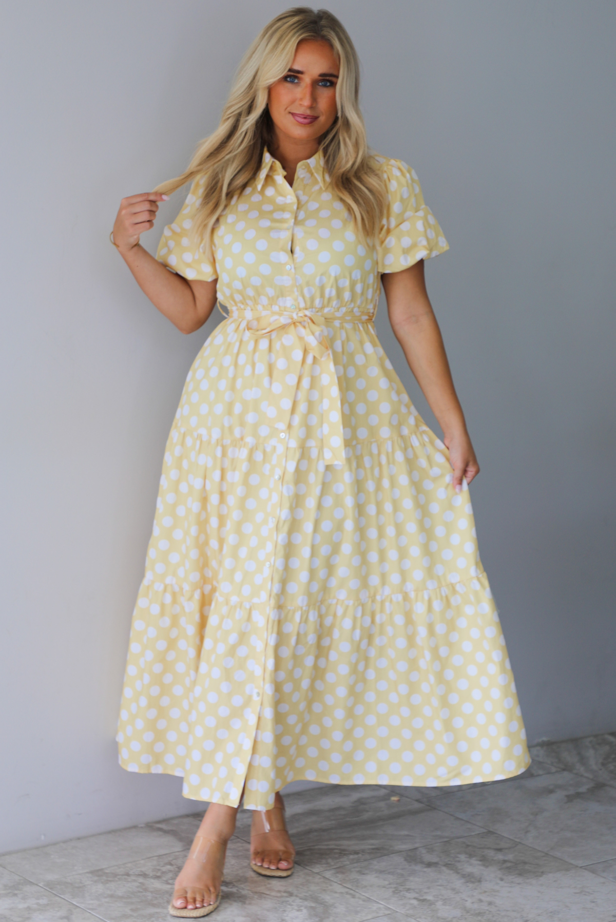 A woman wearing a yellow and white polka dot midi dress with puff sleeves and a tie waist and functional button closures along the front, standing indoors against a white wall with white marble flooring. 