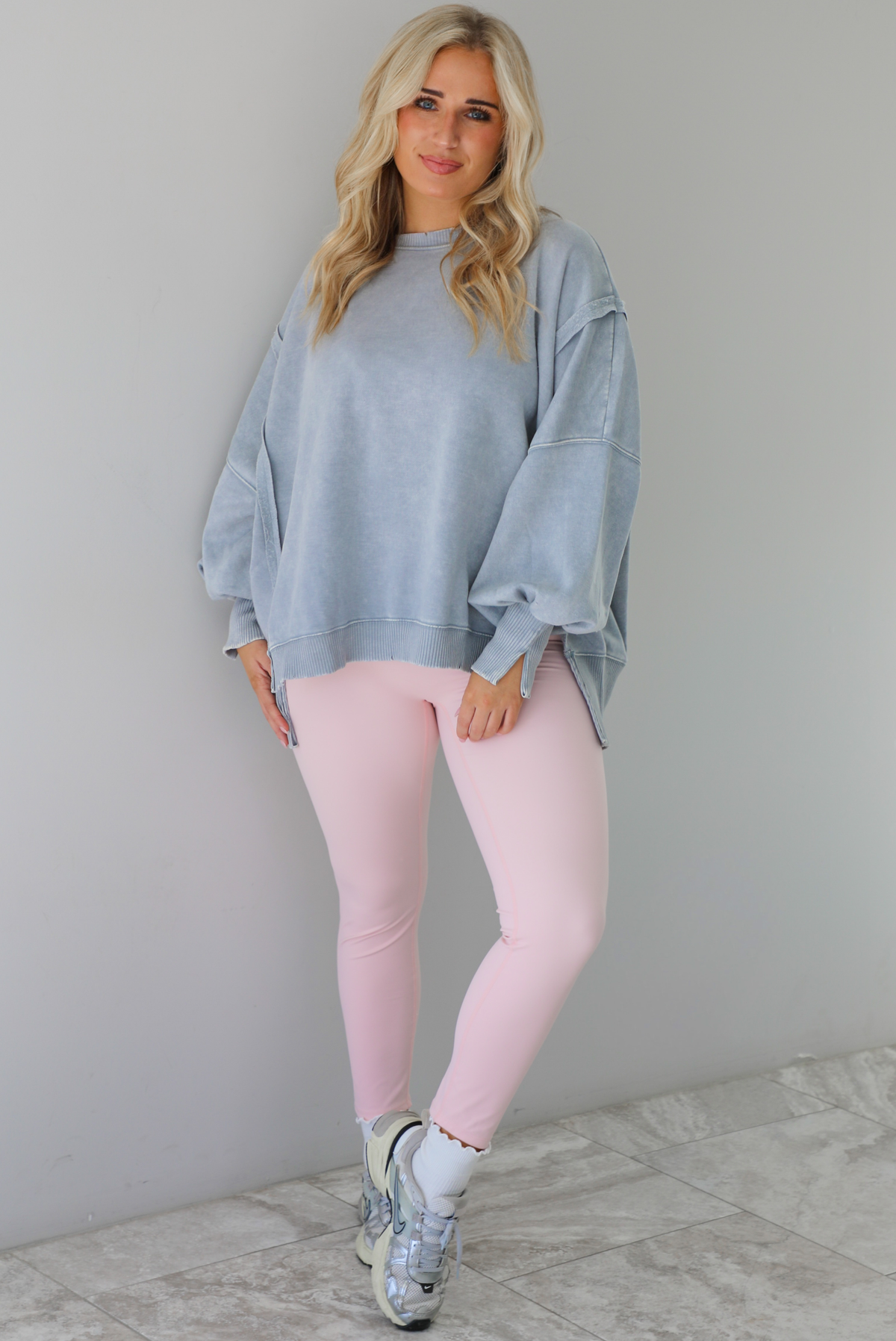 Woman wearing a grey colored oversized sweatshirt with exposed seam details and a high-low design with baby pink leggings. The outfit is styled with white ankle socks and silver sneakers. The photo is taken indoors against a white wall with white marble flooring. 