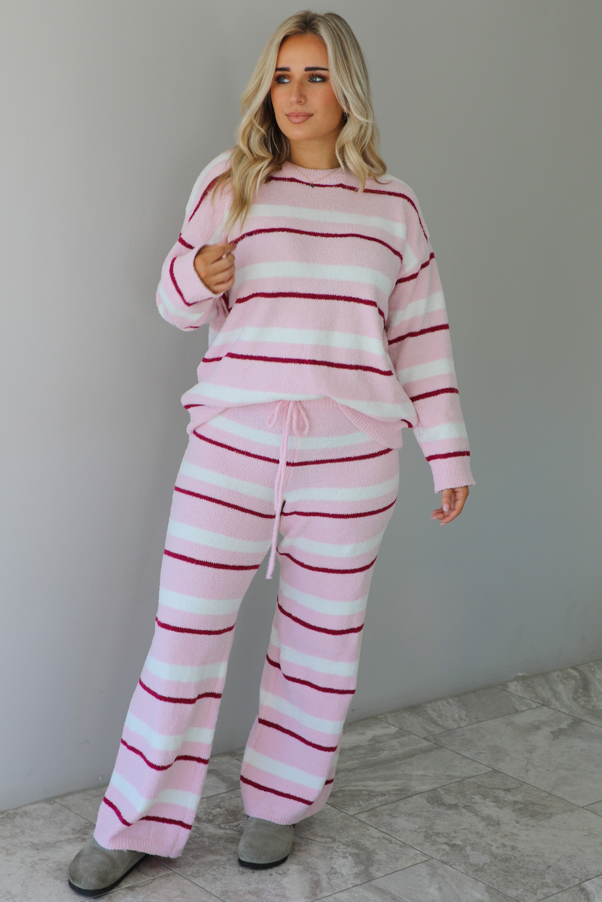 Woman wearing a pink set with a darker pink and white striped design. The set consists of an oversized sweater top and high waisted pants. The sweater of the set is tucked slightly in the front of the pants. The photo is taken indoors against a white wall with white marble flooring. 