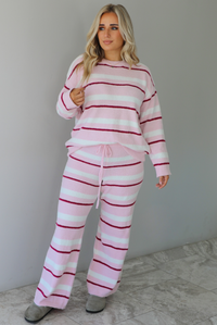 Woman wearing a pink set with a darker pink and white striped design. The set consists of an oversized sweater top and high waisted pants. The sweater of the set is tucked slightly in the front of the pants. The photo is taken indoors against a white wall with white marble flooring. 