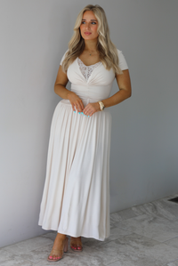 Woman wearing a light sand colored maxi length dress that has a fitted bodice with lace detailing in the center of the bust, a drop waist design, short sleeves, and a flowy skirt. The photo is taken indoors against a white wall with white marble flooring. 