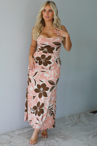 A woman modeling a thick strapped maxi dress with a pink, brown, and white floral pattern, standing indoors against a white wall with white marble flooring. 